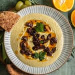 A bowl of white bean dip topped with olives and fresh herbs with fresh fruit in the background.