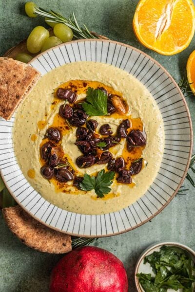 A bowl of white bean dip topped with olives and fresh herbs with fruit and bread in the background.