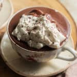 A cup of hot chocolate topped with whipped cream on a saucer over a wooden board.