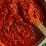 A dutch oven with a chunky marinara sauce, a wooden spoon, and bits of cooked basil.