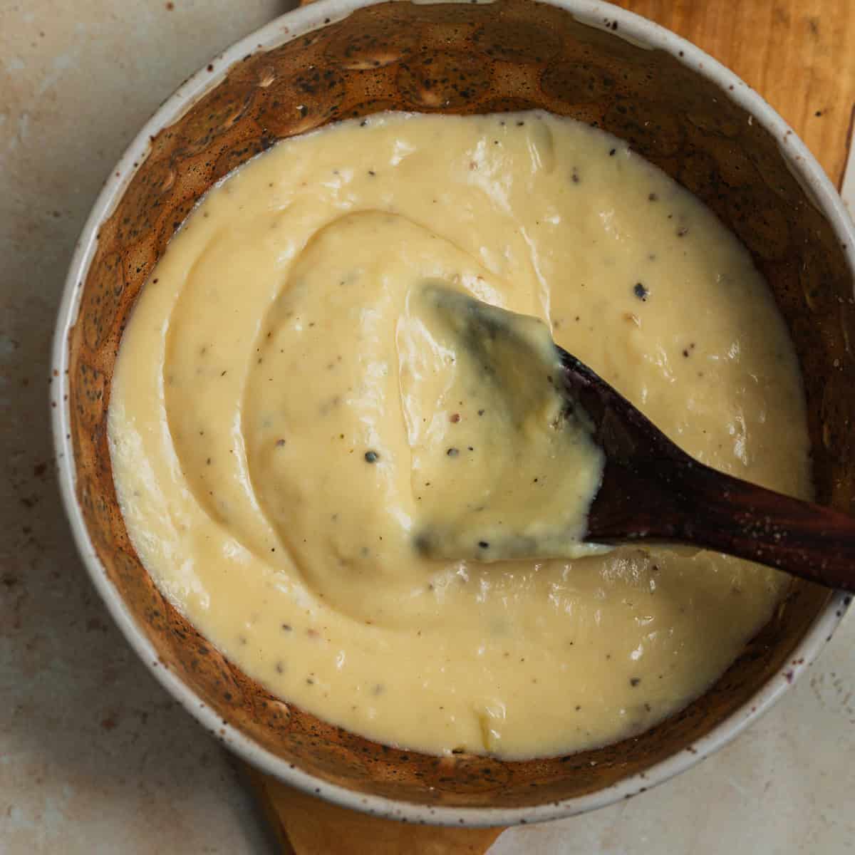 A bowl with a thick, pale yellow, creamy sauce dotted with black pepper and wooden spoon in it.