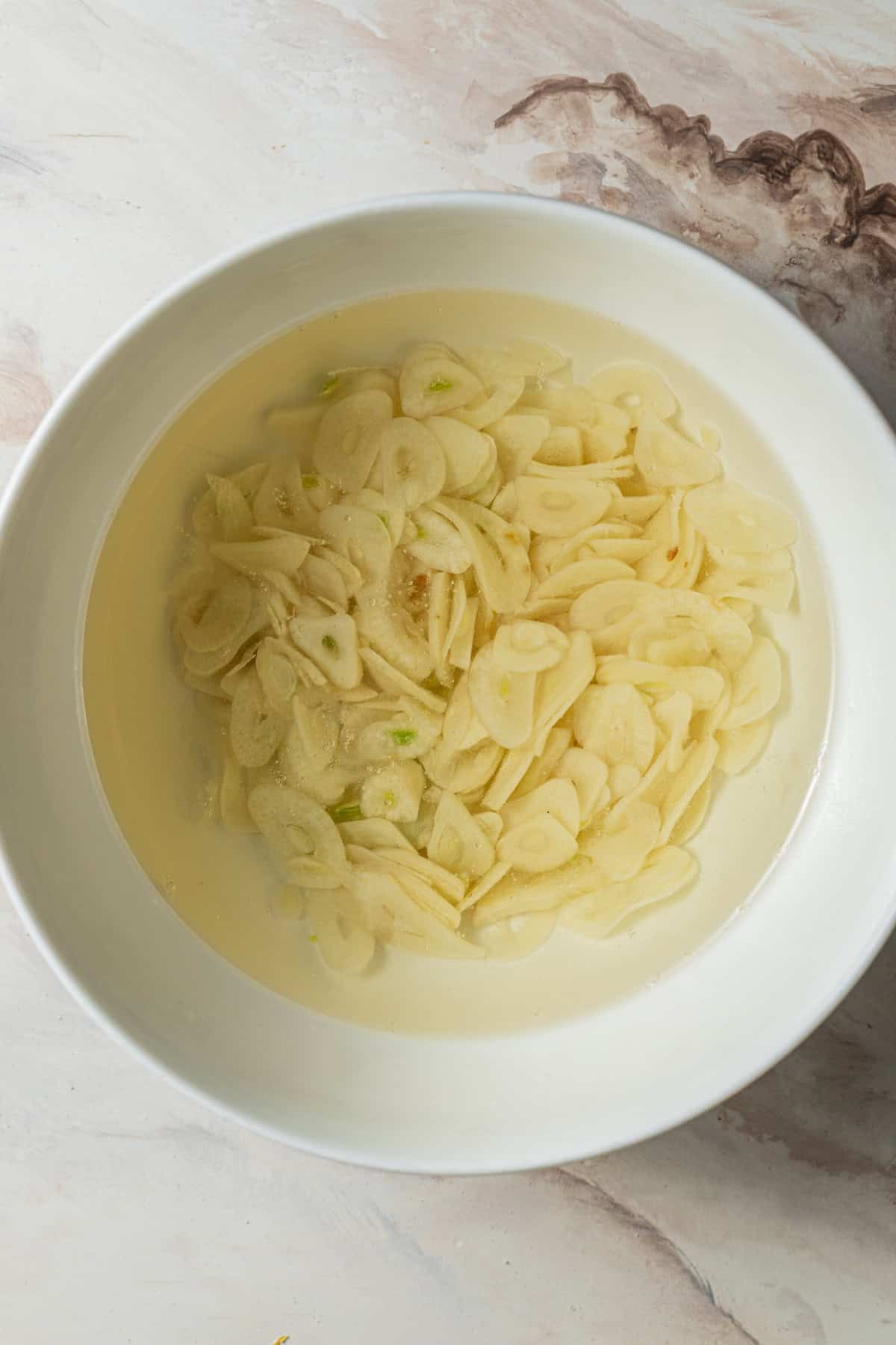 A bowl of thinly sliced garlic submerged in oil on a countertop.