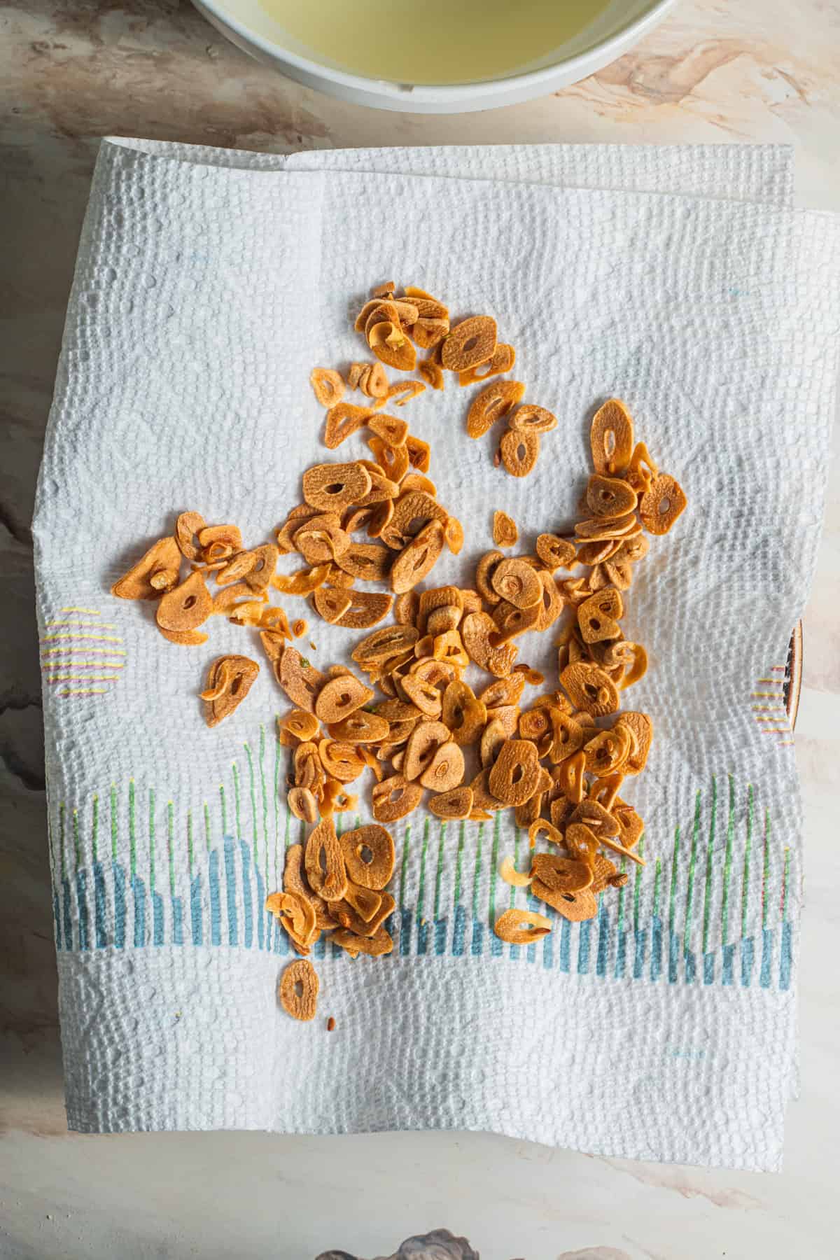 Fried garlic drying on a paper towel with a bowl of oil in the background.