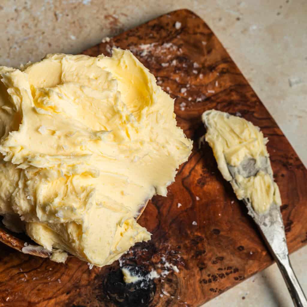 A block of room temperature butter with a buttered knife next to it on a wooden cutting board.