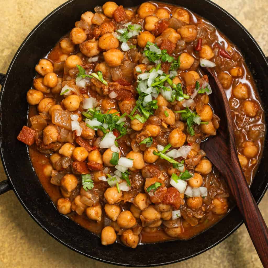A kadai with chickpeas in a tomato sauce topped with onions and fresh cilantro.