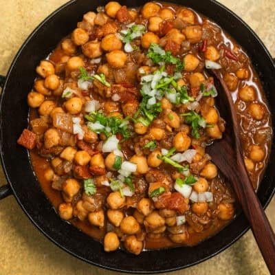 A kadai with chickpeas in a tomato sauce topped with onions and fresh cilantro.