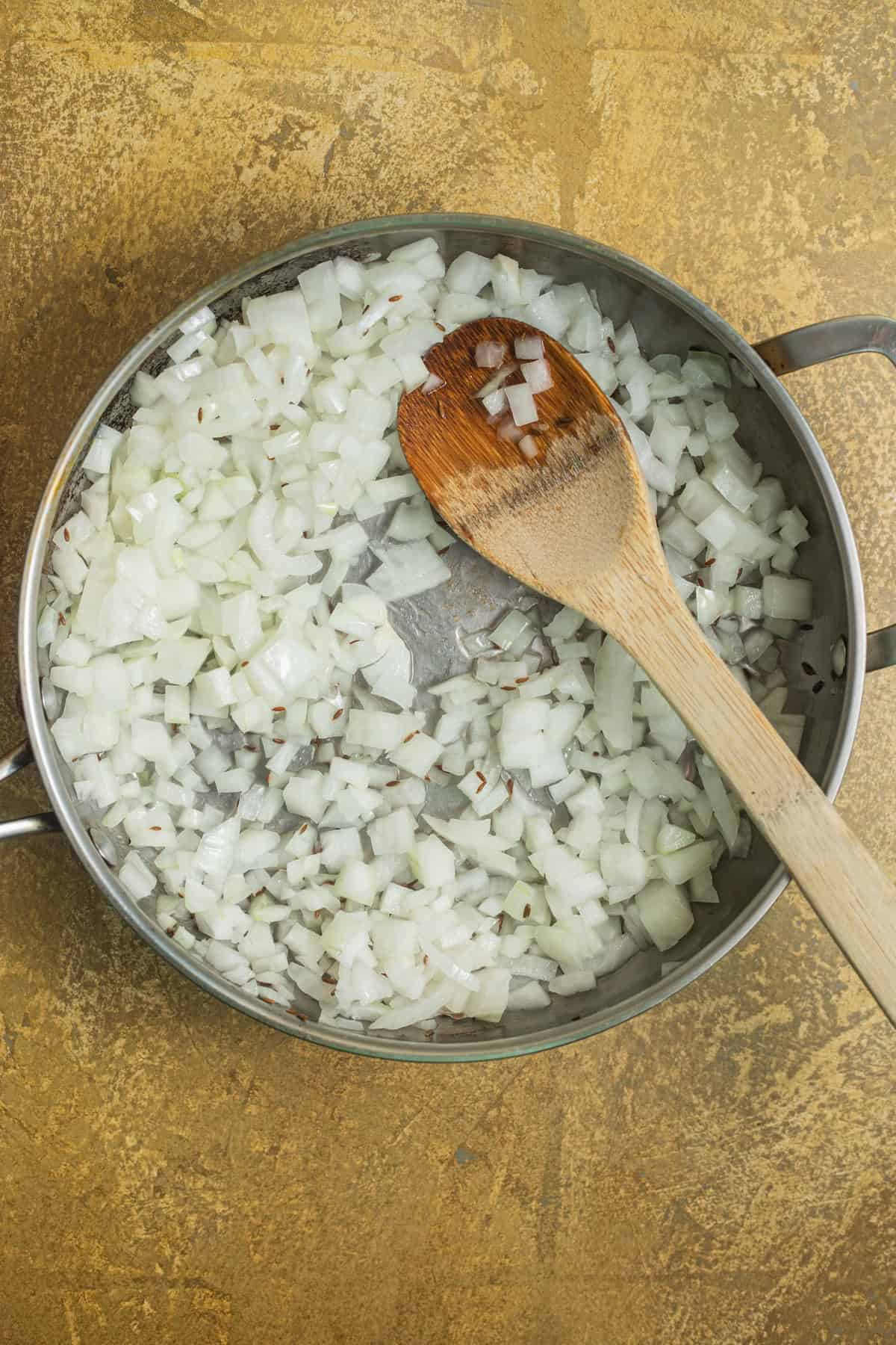 A stainless steel skillet with chopped raw onion and a wooden spoon.