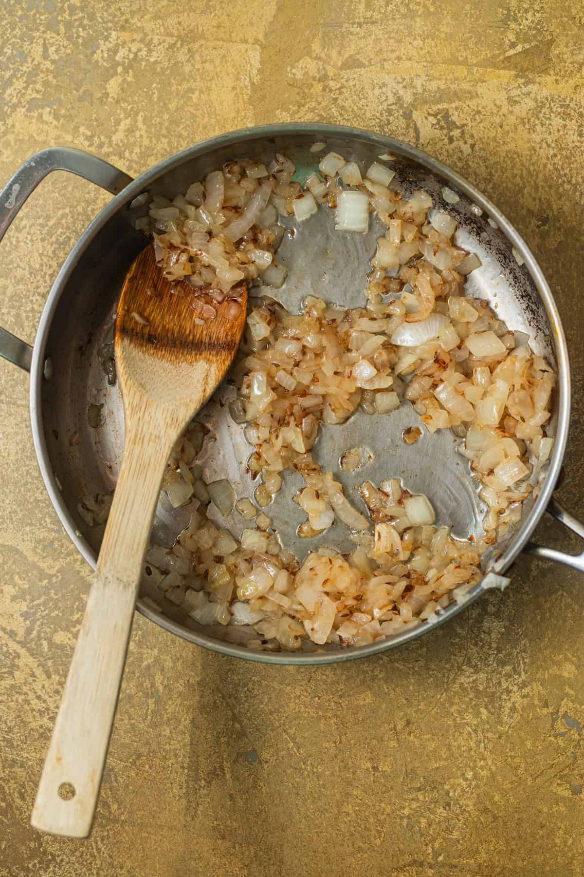 A stainless steel skillet with golden-brown onions sauteeing with a wooden spoon.