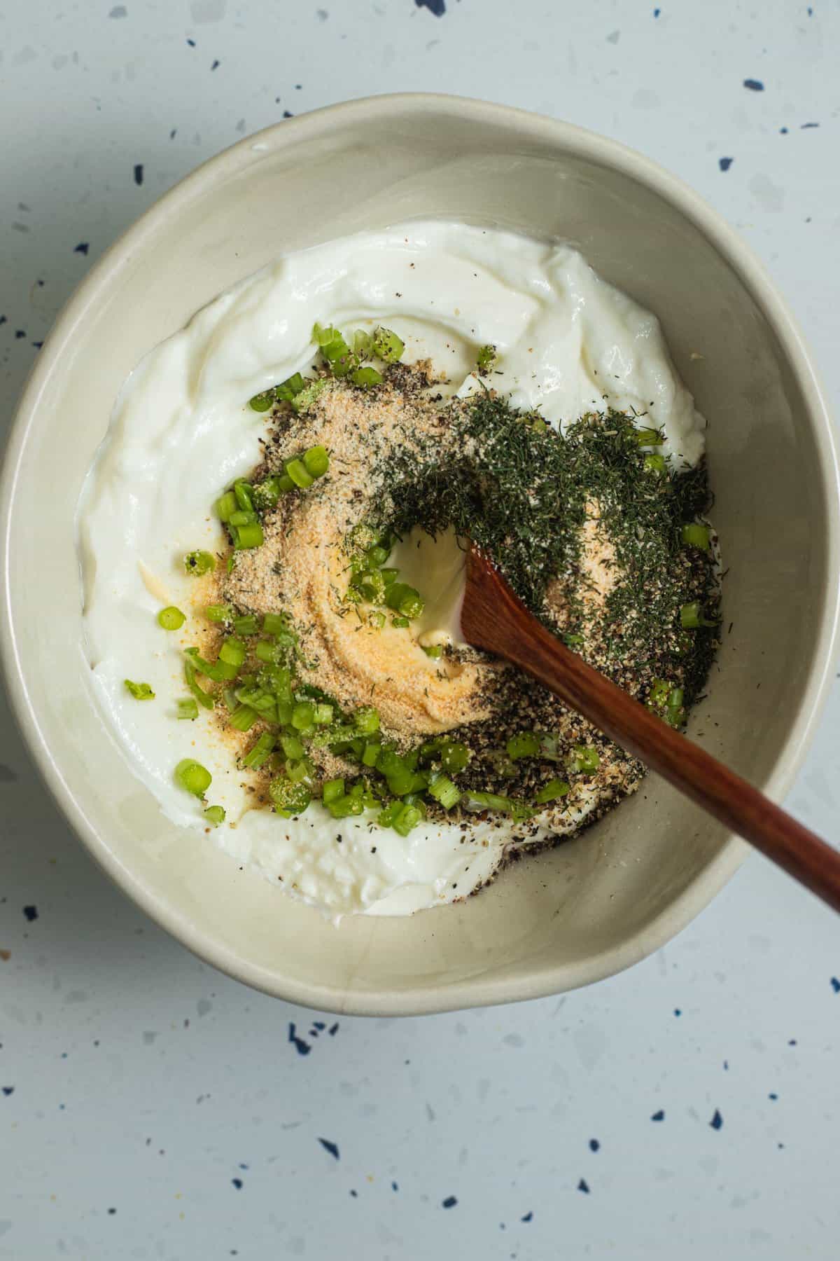 A spoon in a bowl of Greek yogurt with chopped scallions, dried herbs, and spices.