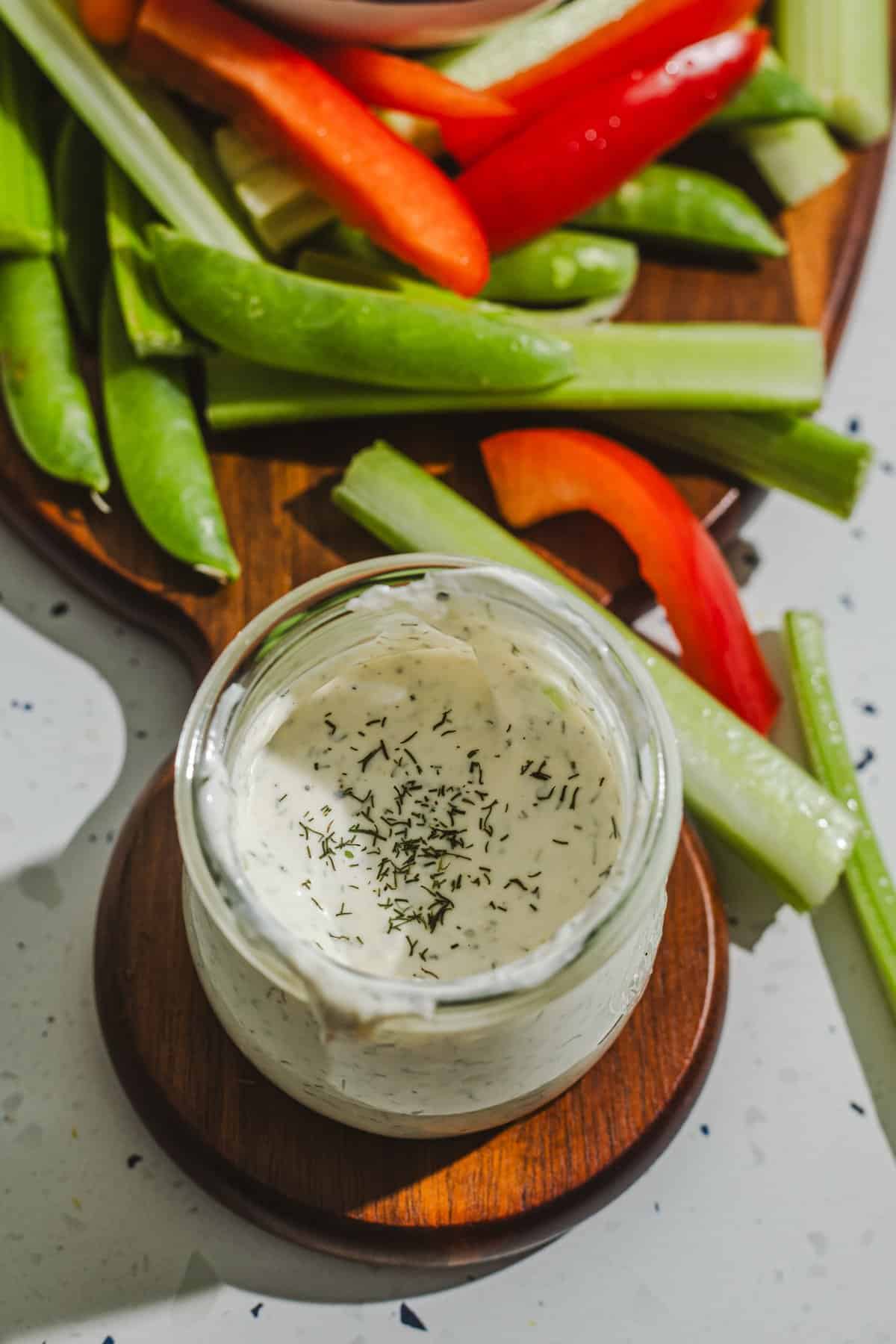 A small jar with ranch dressing and crudites on a wooden cutting board in the background.