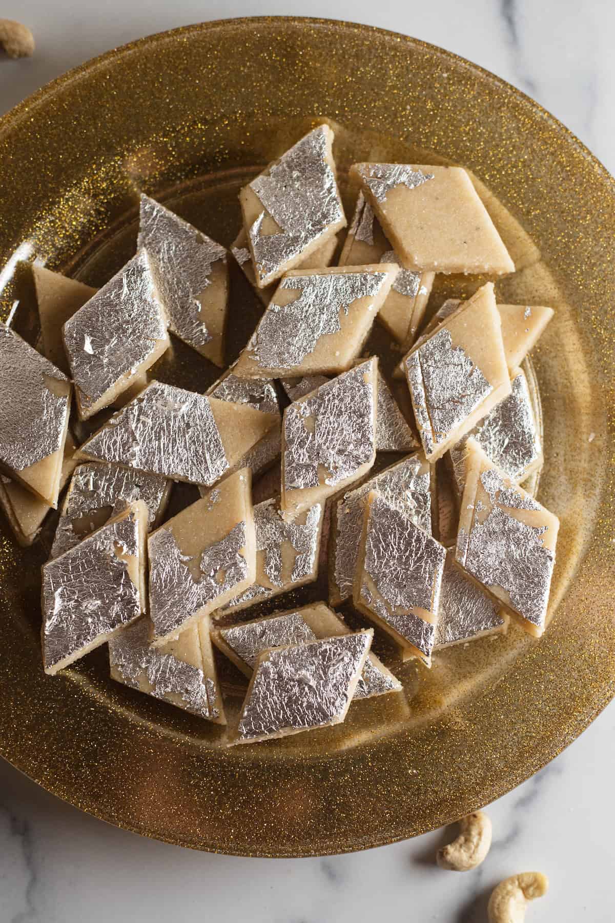 Silver vark decorated kaju katli pieces on a shimmery gold plate with cashews in the background.