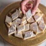 Hand holding a piece of kaju katli over a plate of kaju katli.