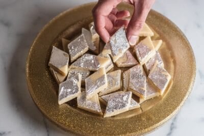 Hand holding a piece of kaju katli over a plate of kaju katli.