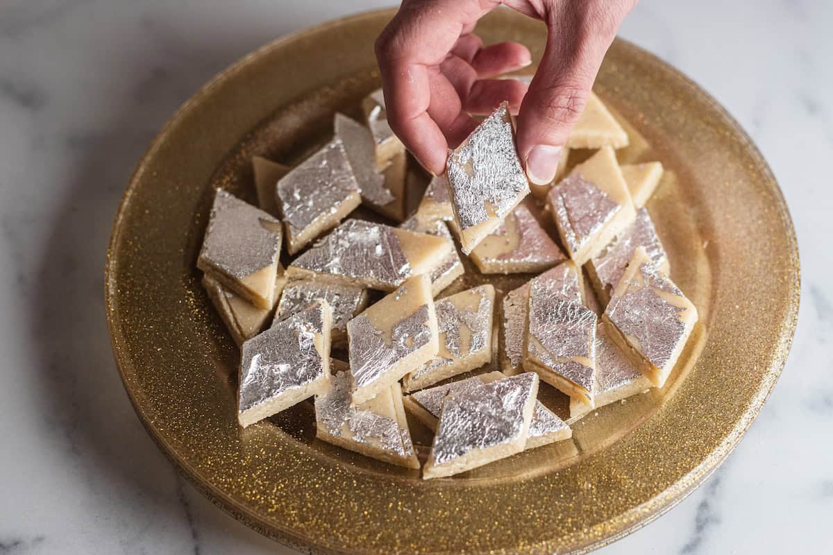 Hand holding a piece of kaju katli over a plate of kaju katli.