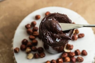 Knife scooping chocolate spread out of a jar surrounded by hazelnuts.