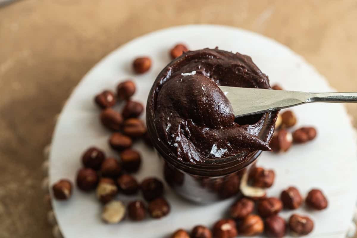 Knife scooping chocolate spread out of a jar surrounded by hazelnuts.