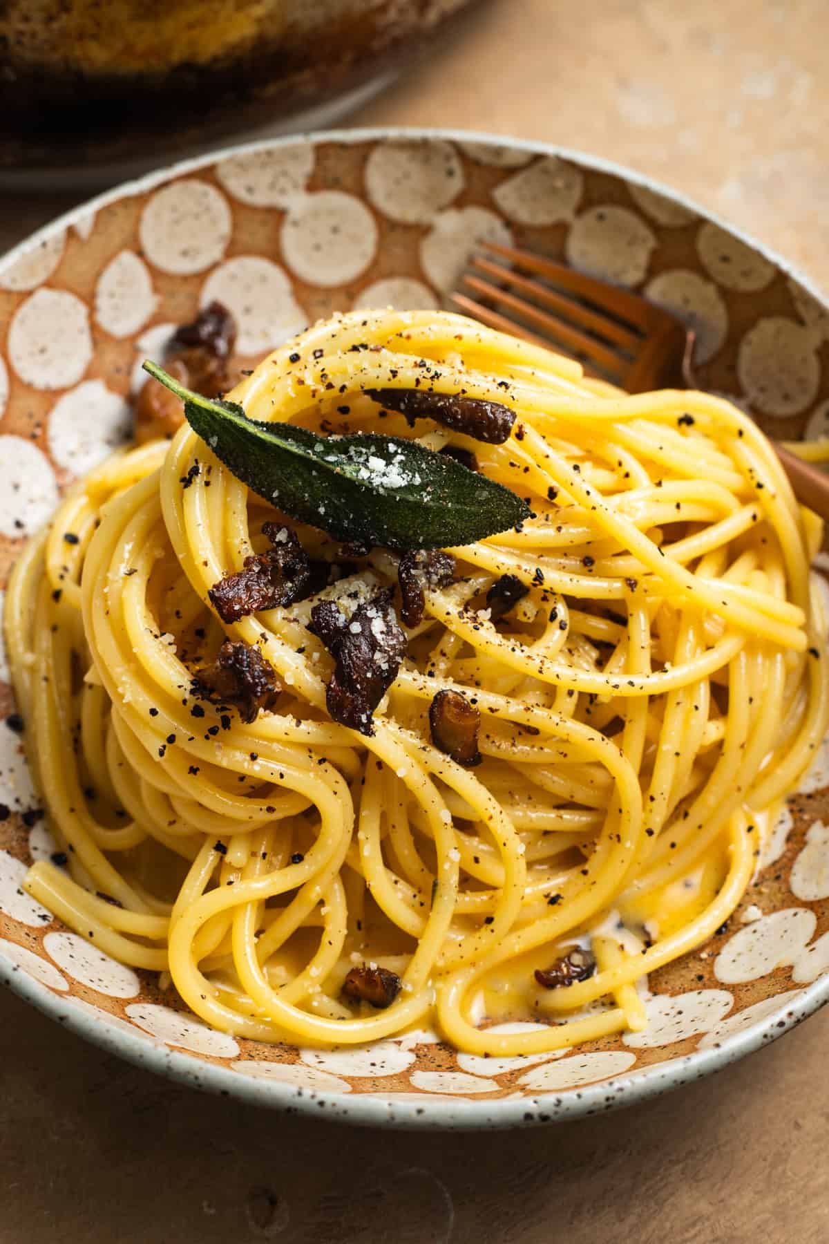 A bowl of spaghetti tossed in carbonara sauce and topped with sage leaves, mushrooms, cheese, and black pepper.