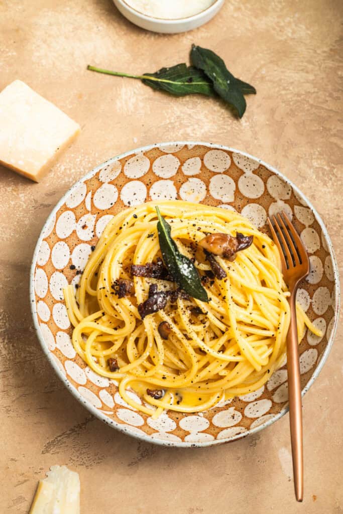 A bowl of spaghetti tossed in carbonara sauce and topped with sage leaves, mushrooms, cheese, and black pepper.