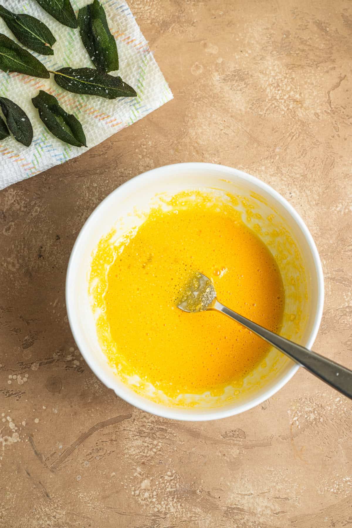 A bowl of whisked egg yolks with a plate of sage leaves in the background.
