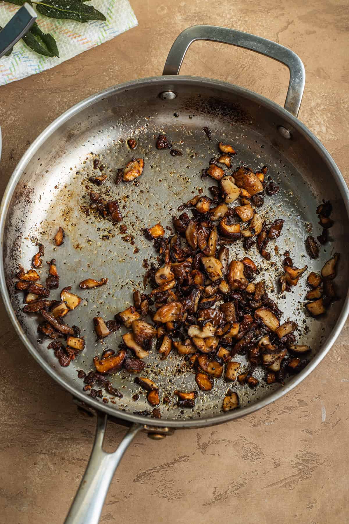 A stainless steel skillet with crisped up mushrooms and sage leaves on a paper towel in the background.