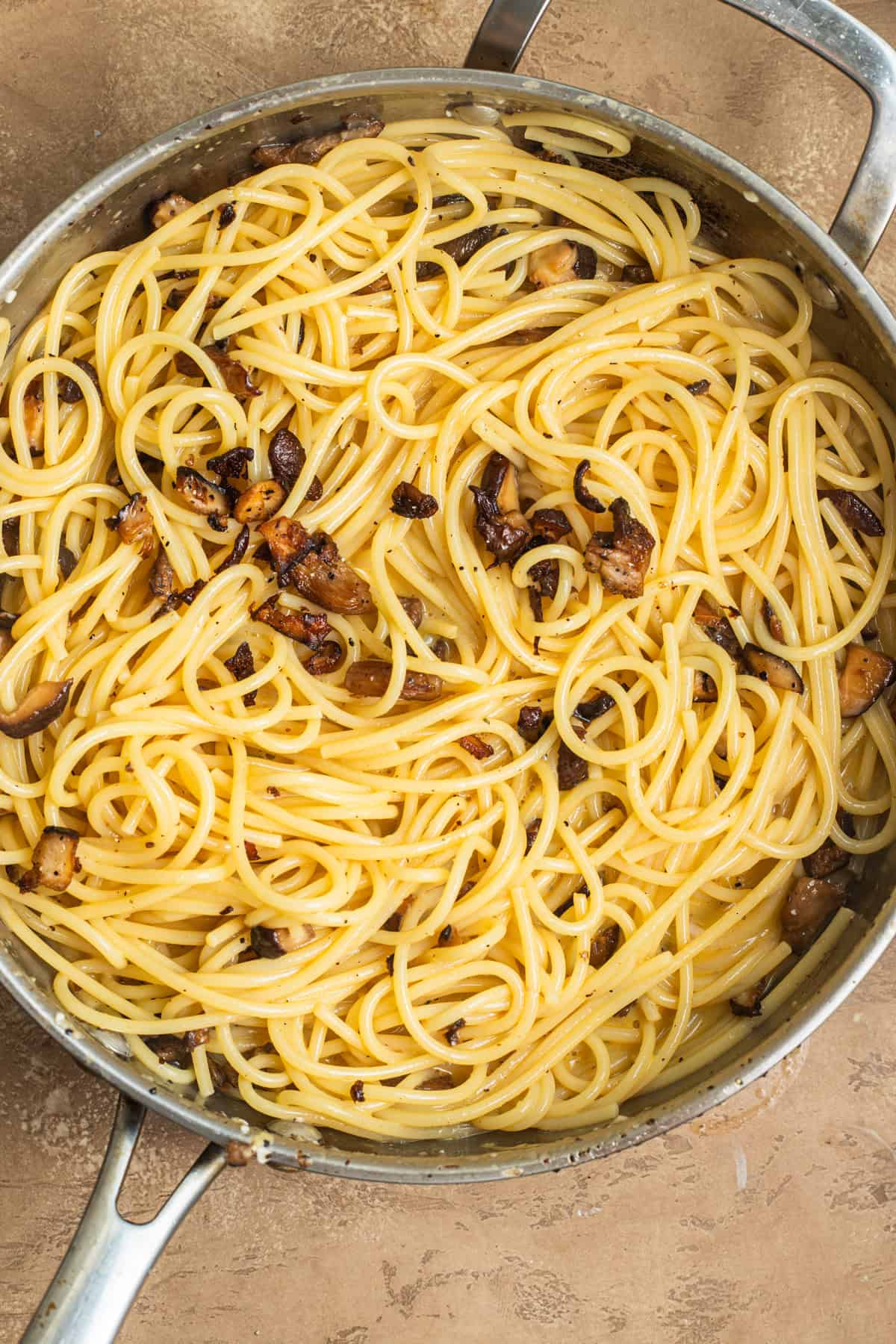 Spaghetti in a creamy sauce with small bits of mushrooms in a stainless steel skillet.