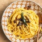 A bowl of pasta tossed in carbonara sauce and topped with sage leaves and mushrooms.