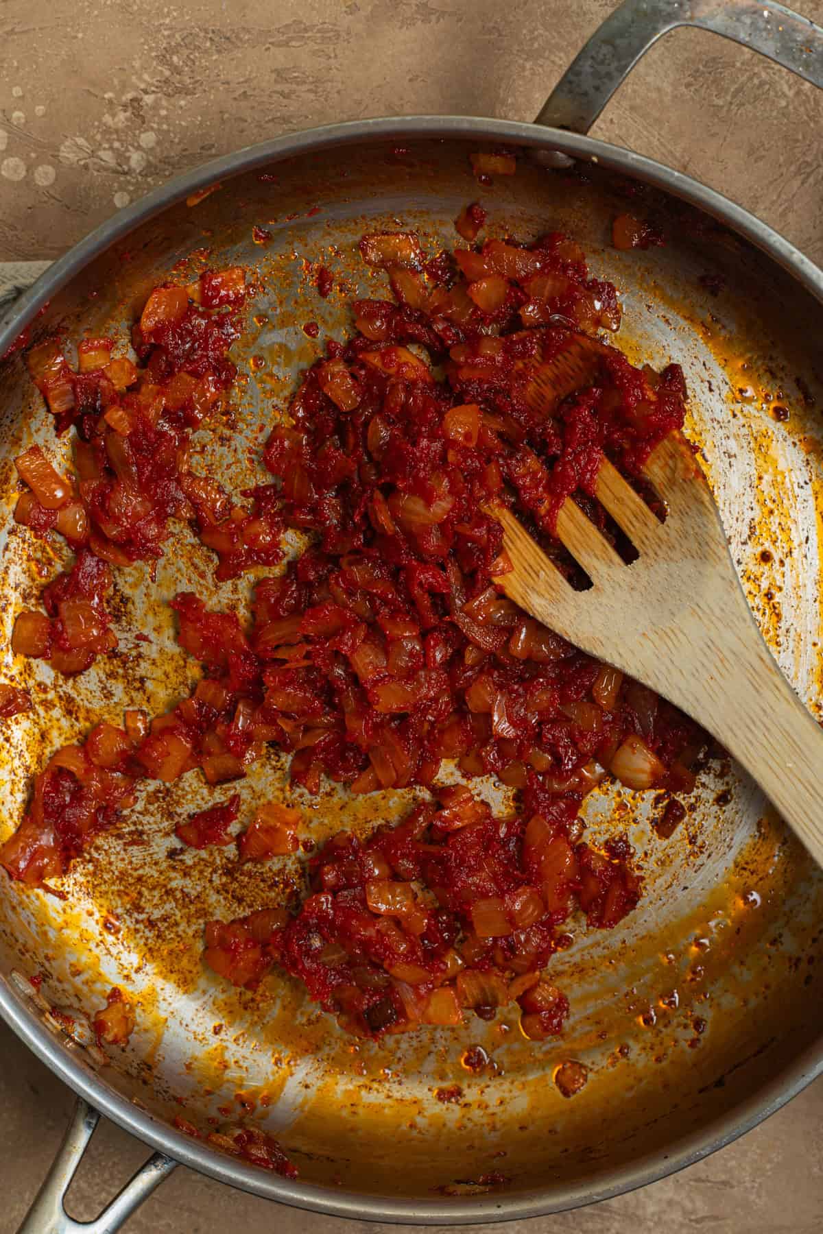 A stainless steel skillet with tomato-paste coated onions with a wooden spoon.