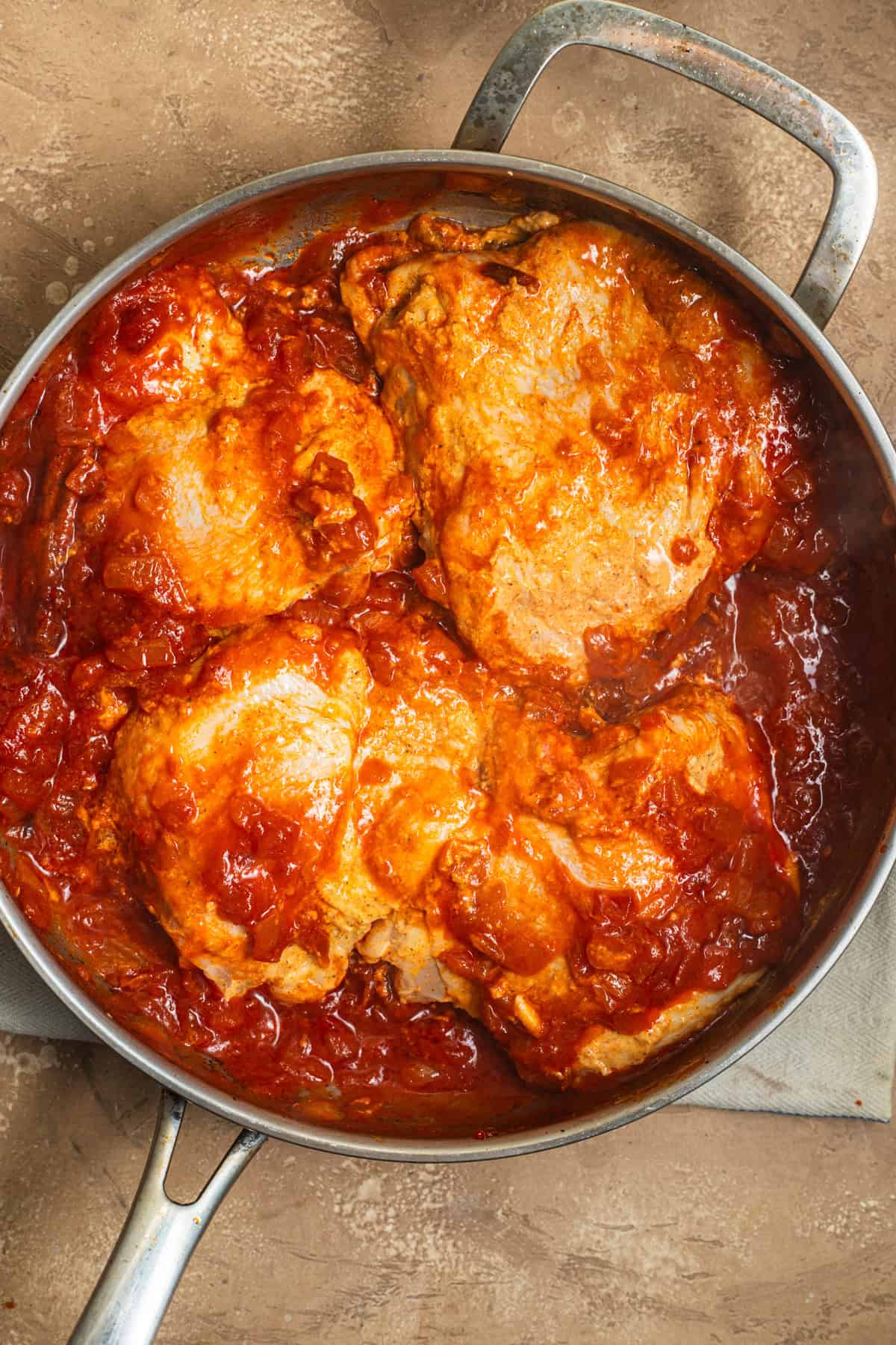 A stainless steel skillet with chicken thighs nestled in a tomato sauce.