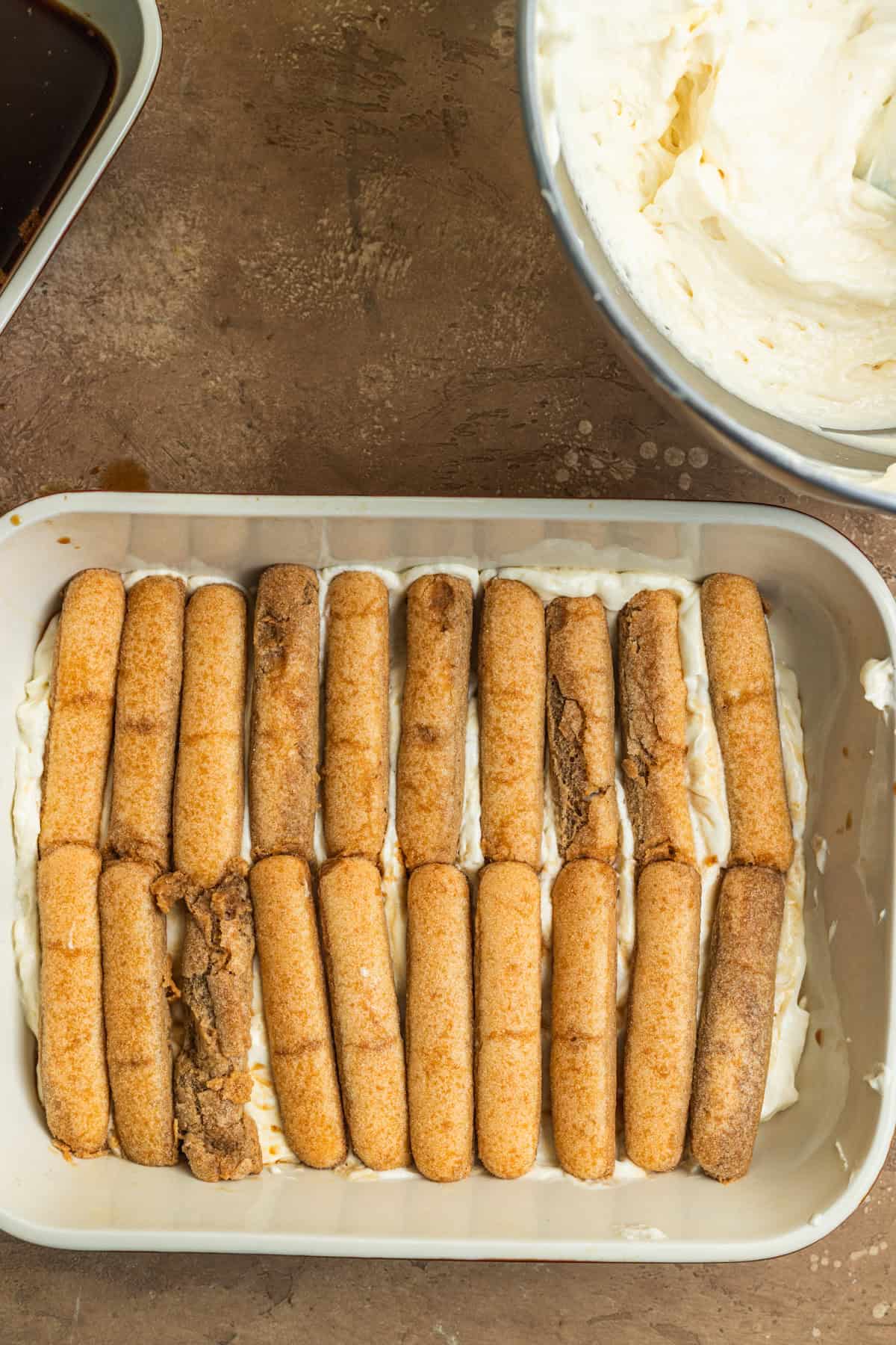 A casserole dish with rows of ladyfingers sitting atop a layer of cream.