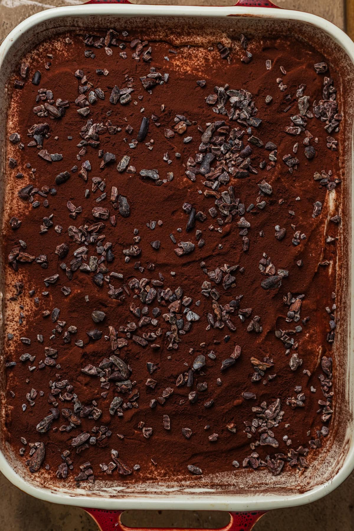 A casserole dish filled with tiramisu topped with cocoa powder and cocoa nibs.