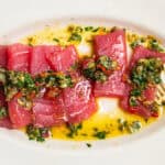 A plate of tuna crudo topped with a caper herb dressing in a pool of olive oil.