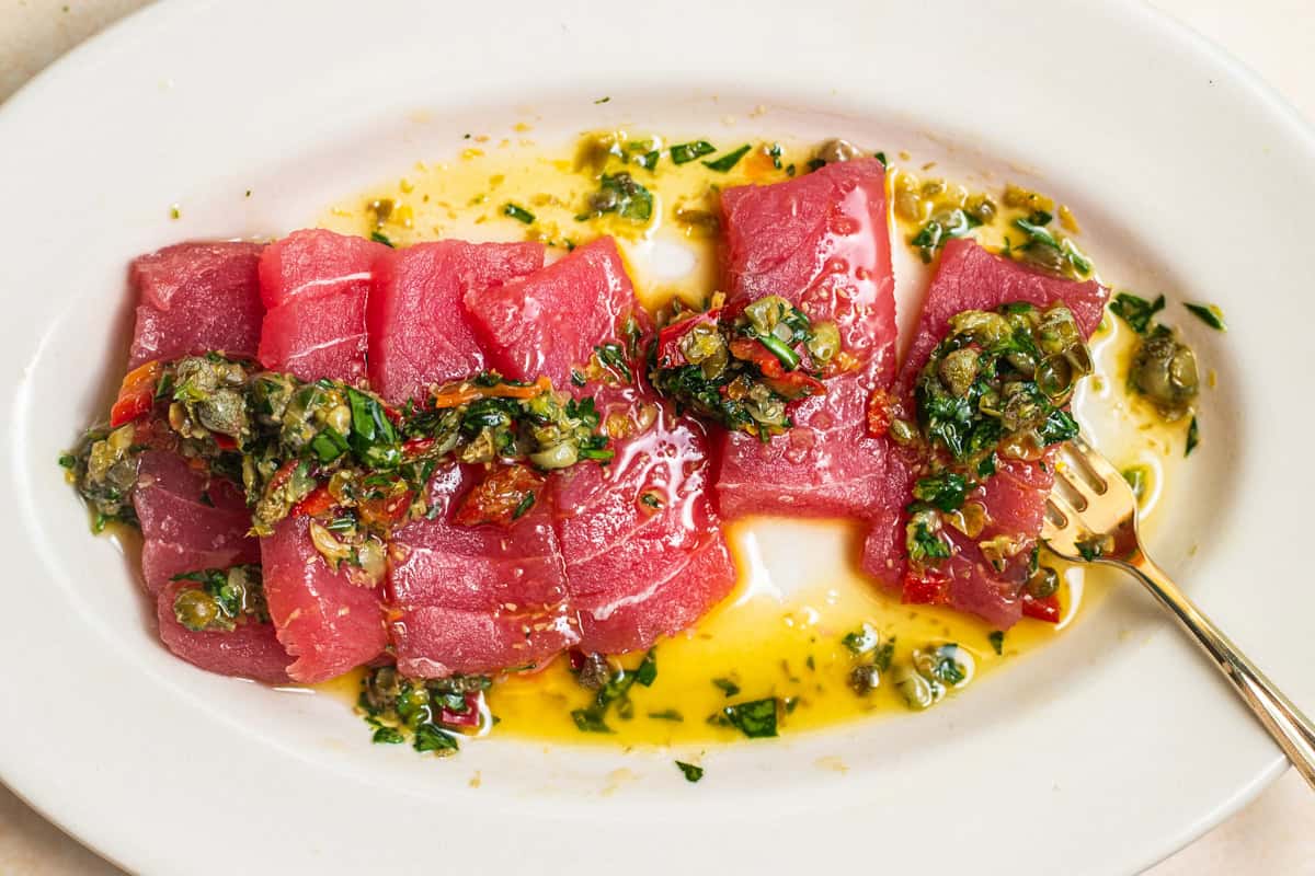 A plate of tuna crudo topped with a caper herb dressing in a pool of olive oil.