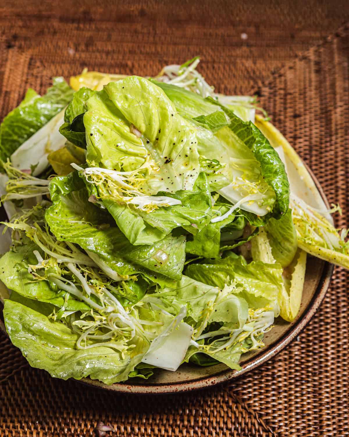 A pile of various lettuces, frisee, and endive set on a plate over a woven placemat. 