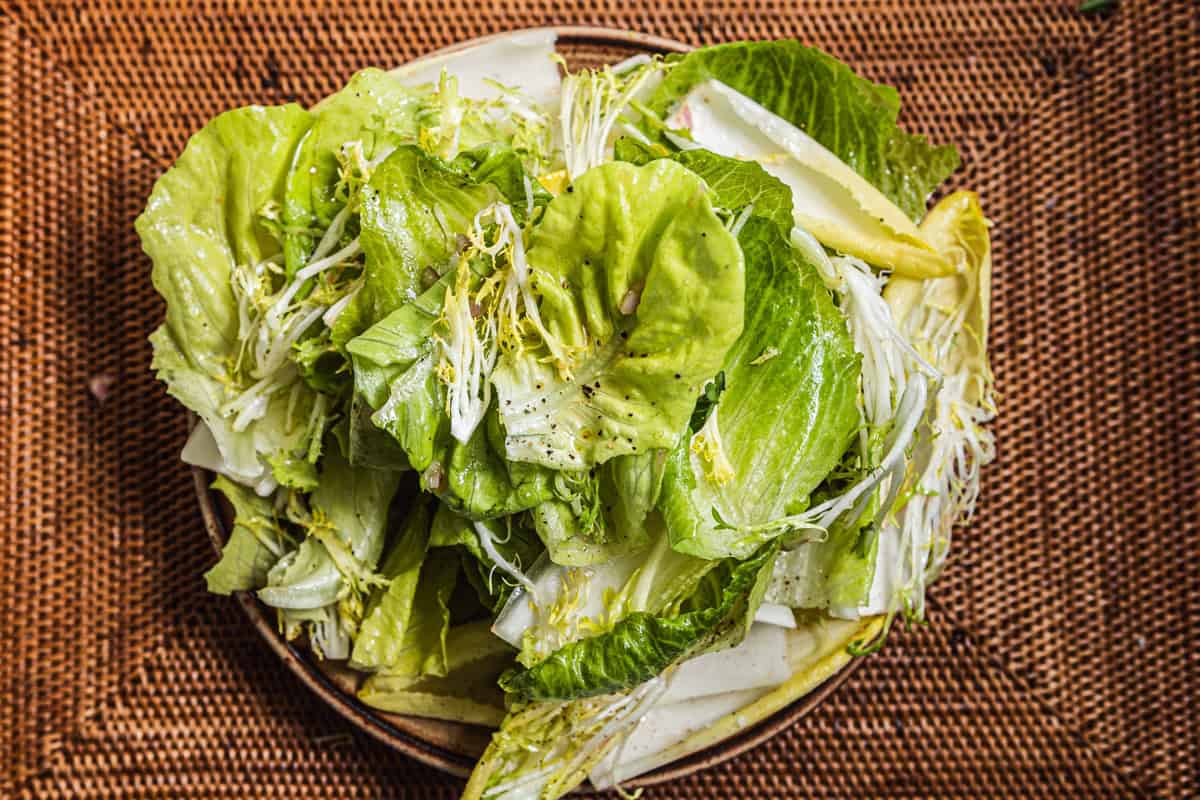 Piles of various lettuces, frisee, and endive dressed with salad dressing on a plate set over a woven placemat.