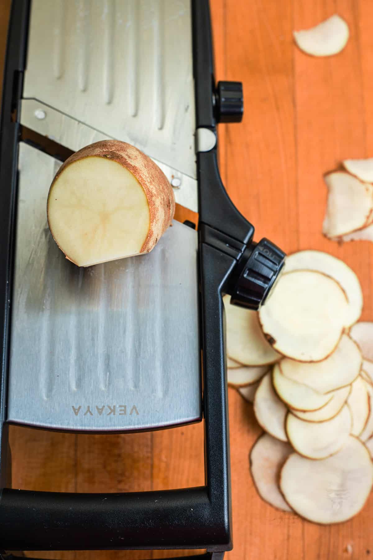 A potato being sliced on a mandoline set over a cutting board.