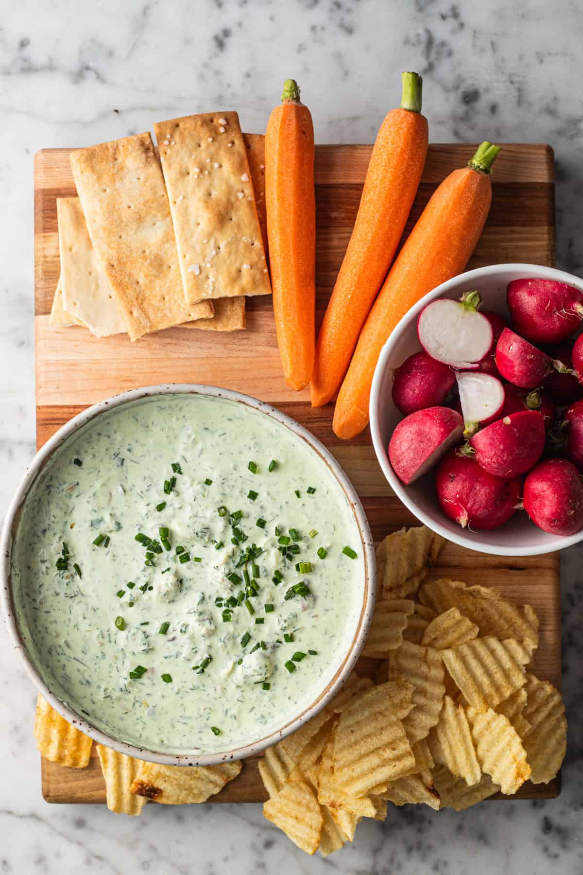 Crudites, crackers, and chips on a wooden cutting board with a bowl of smoked trout dip next to it.