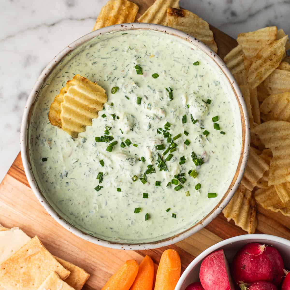 A bowl of pale green smoked trout dip topped with chives with a chip inside on a platter of crudite.
