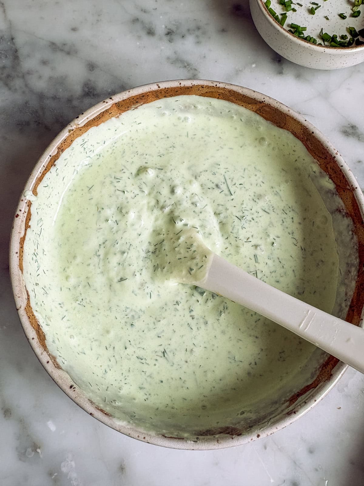 A bowl of sour cream and creme fraiche blended with fresh herbs and a bowl of chives in the background.