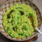 A bowl of green pea risotto topped with green peas and brown butter with a spoon.