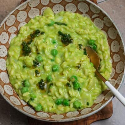 A bowl of green pea risotto topped with green peas and brown butter with a spoon.
