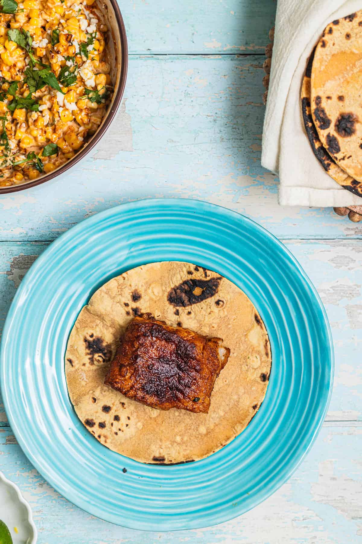 A plate with a fish taco with a bowl of corn and plate of tortillas in the background.