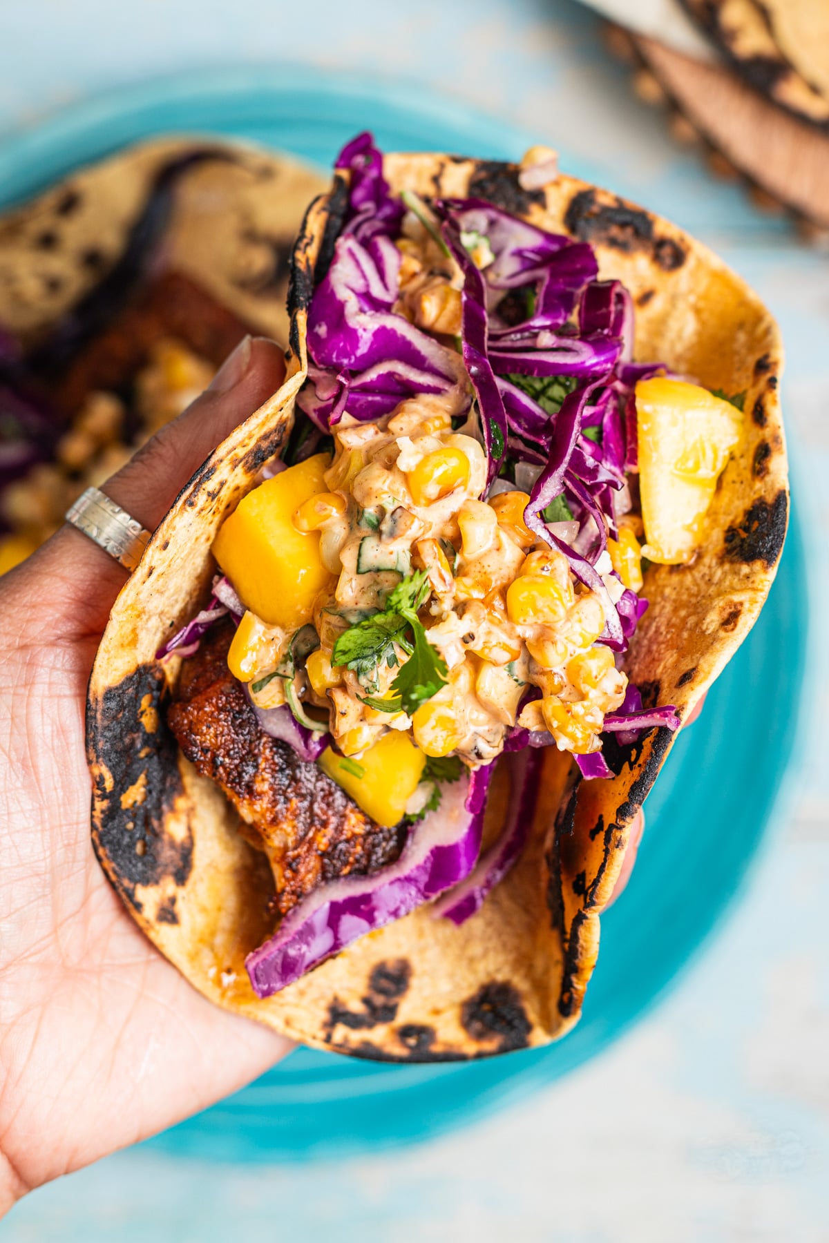 Hand holding a charred tortilla taco with fish, mango, cabbage, and a creamy corn salsa.