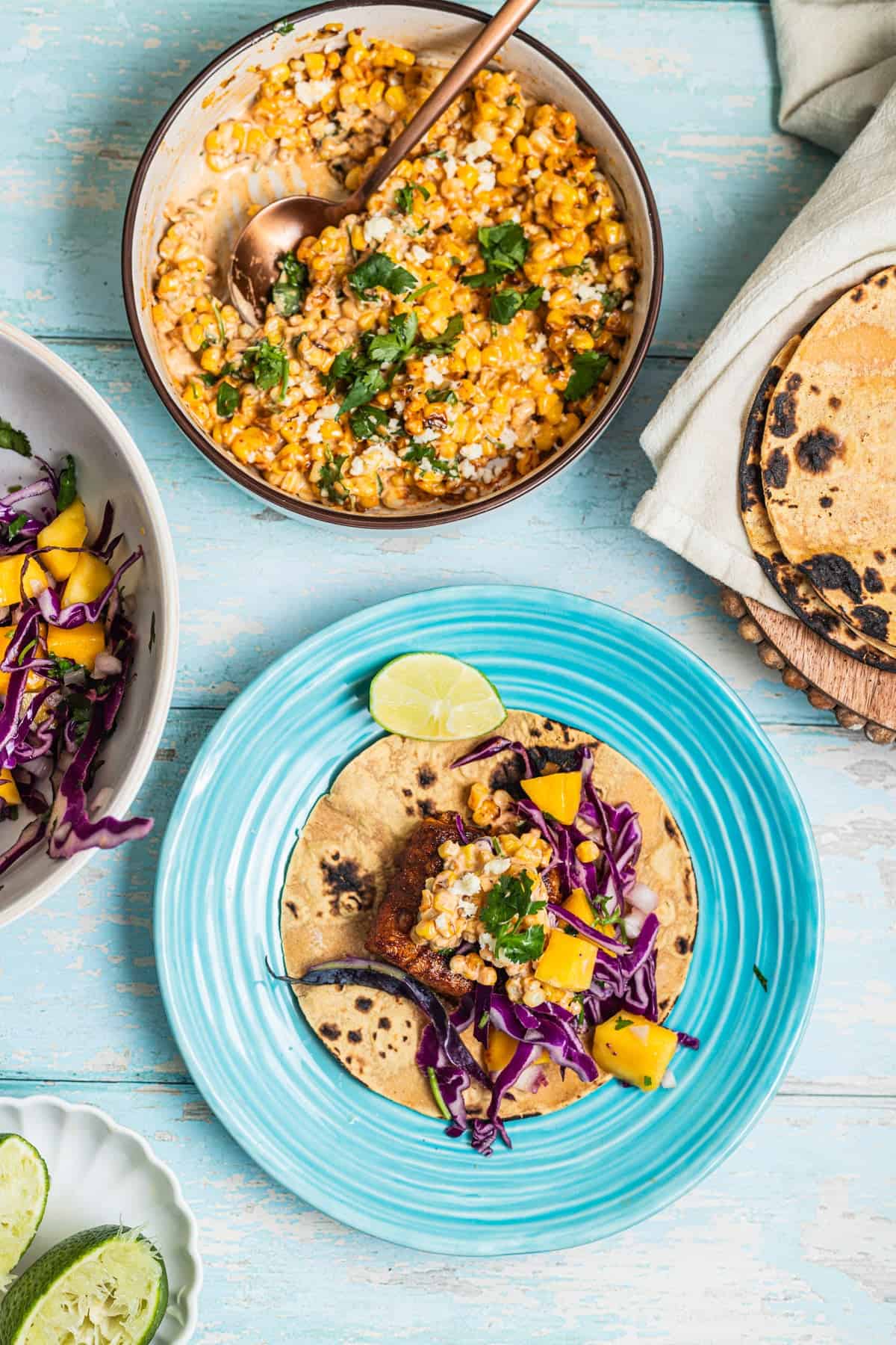 A plate with a fish taco with a mango slaw, corn salad, and charred tortillas in the background.