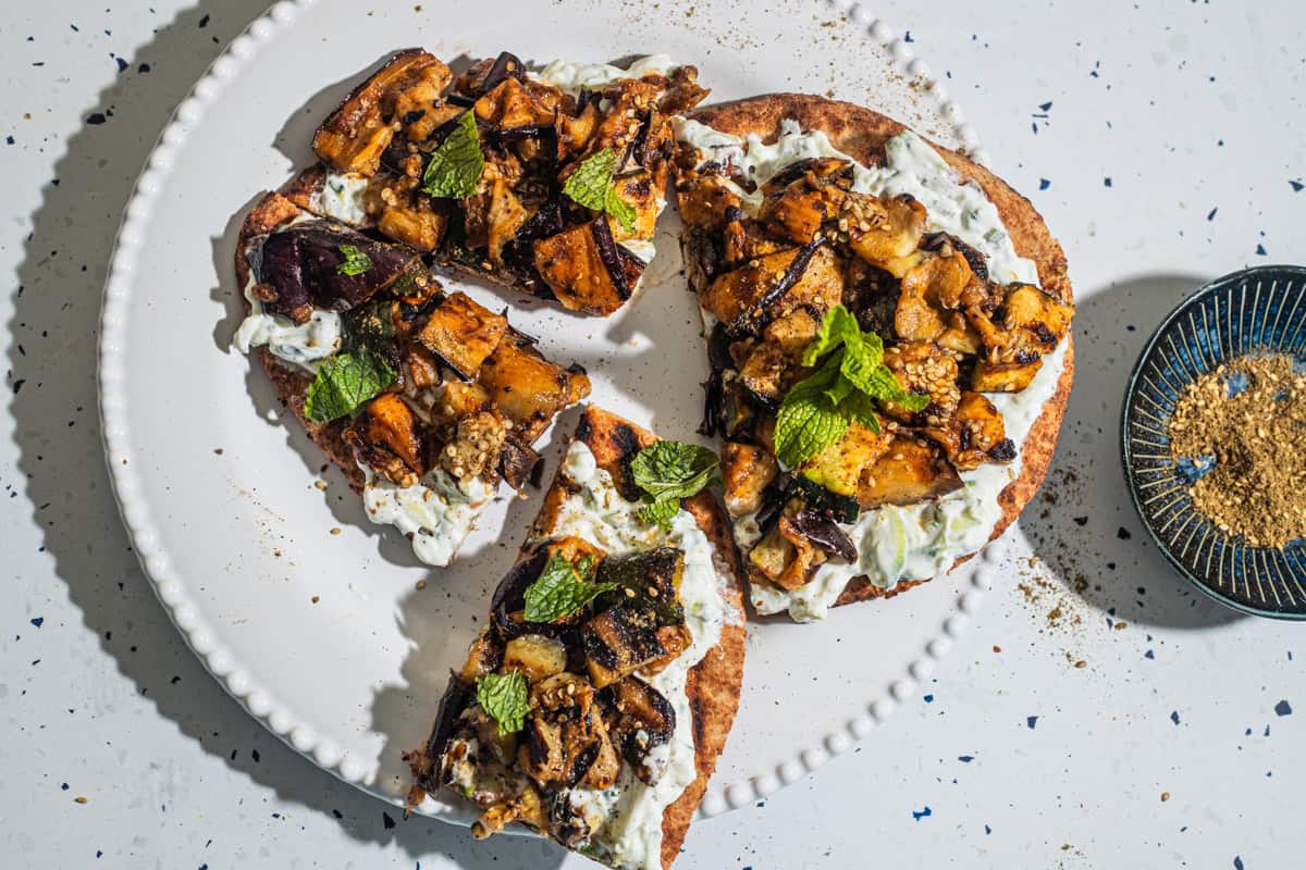 Flatbread pieces with a yogurt sauce and vegetables on a plate with a bowl of za'atar in the background.