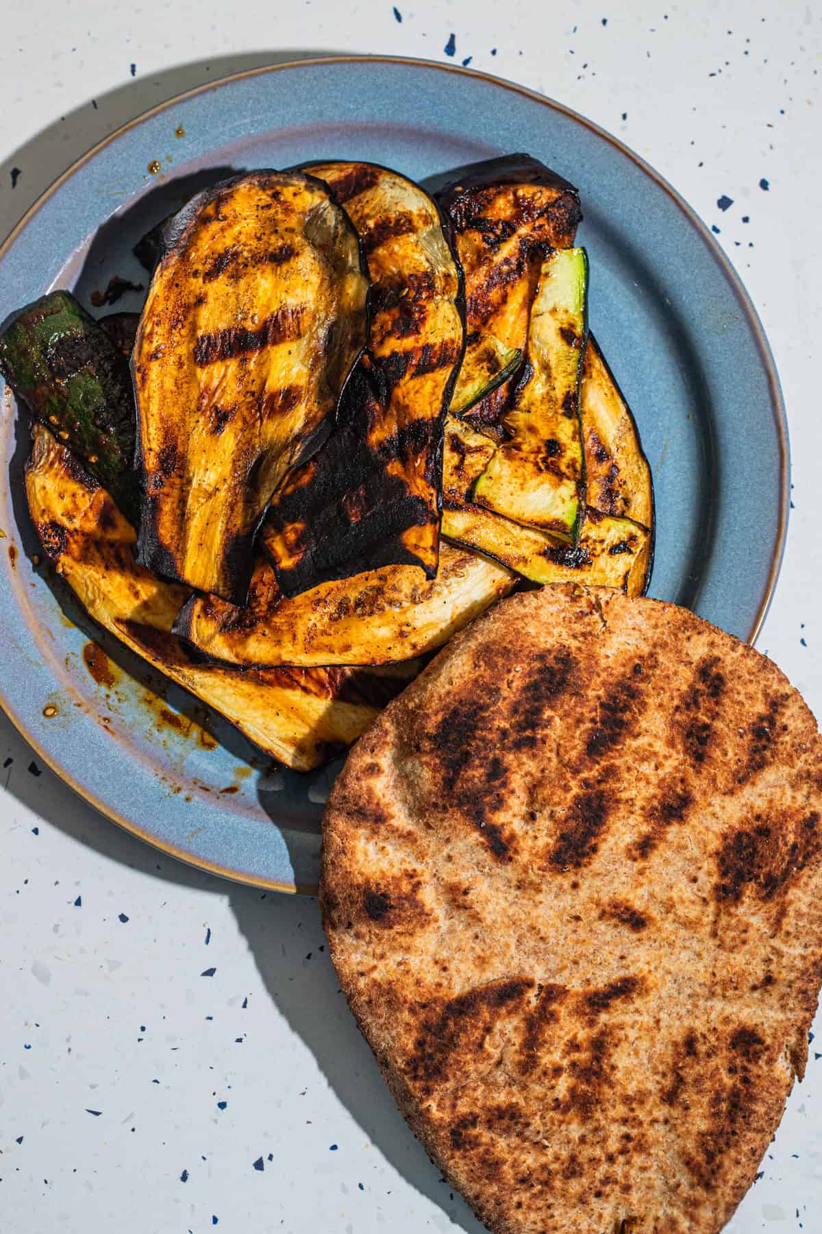 A plate of grilled vegetables with a piece of grilled naan in the background.