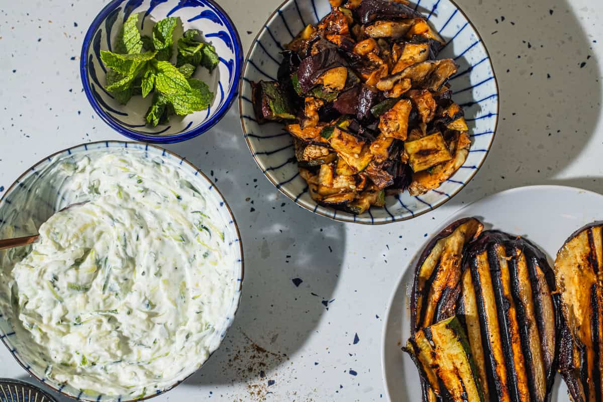 A bowl of mint, a bowl of grilled vegetables, a plate of grilled eggplant, and a bowl of tzatizki.