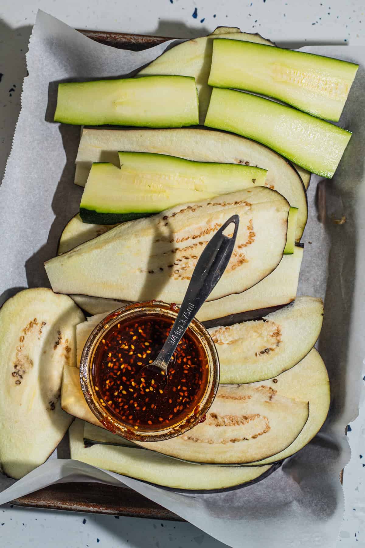 Thinly sliced zucchini and eggplant with a bowl of sauce on a baking sheet.