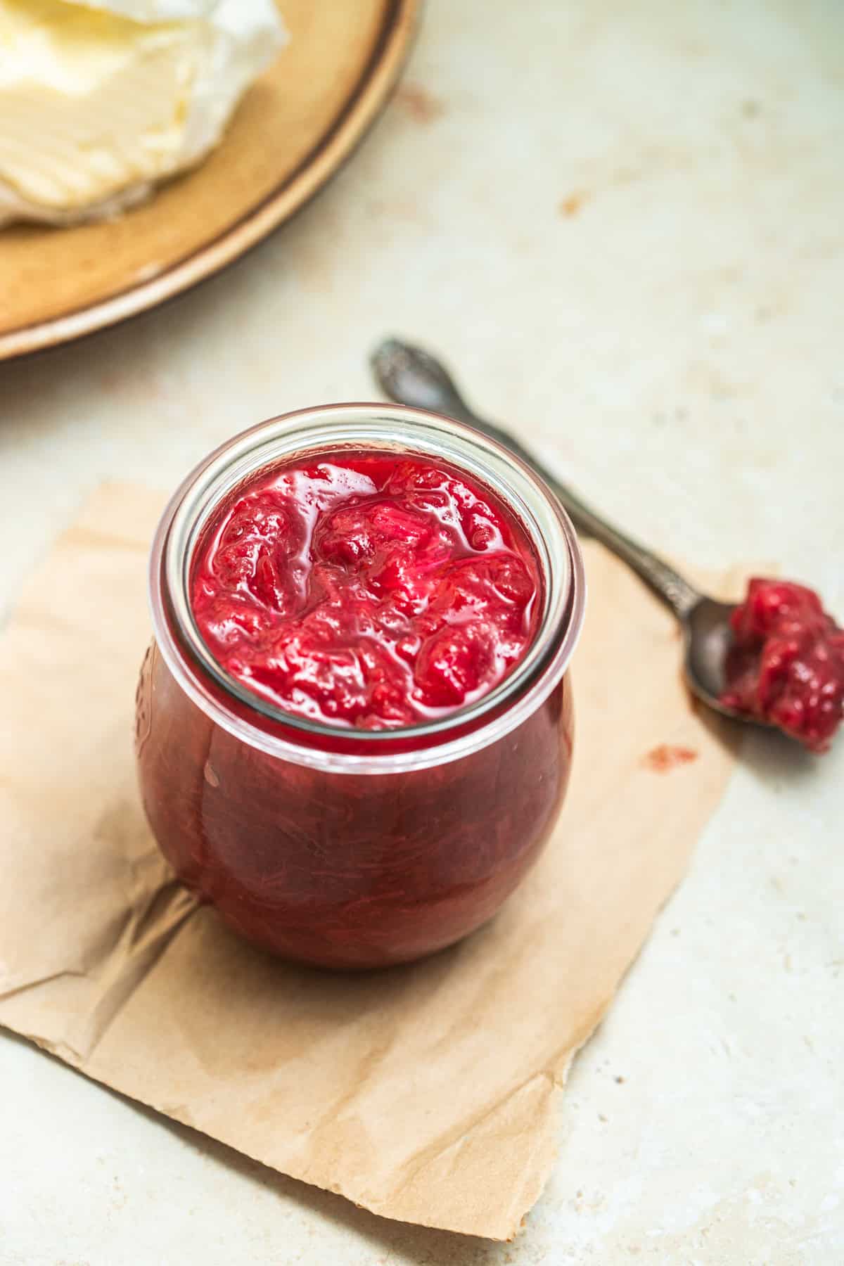 Rhubarb jam in a jar with a spoon of rhubarb and a plate of butter in the background.