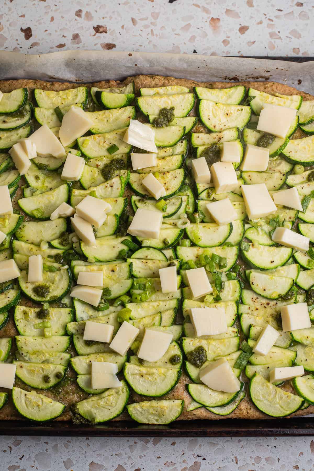 Pizza dough topped with sliced zucchinis, pesto, and mozzarella cubes. 