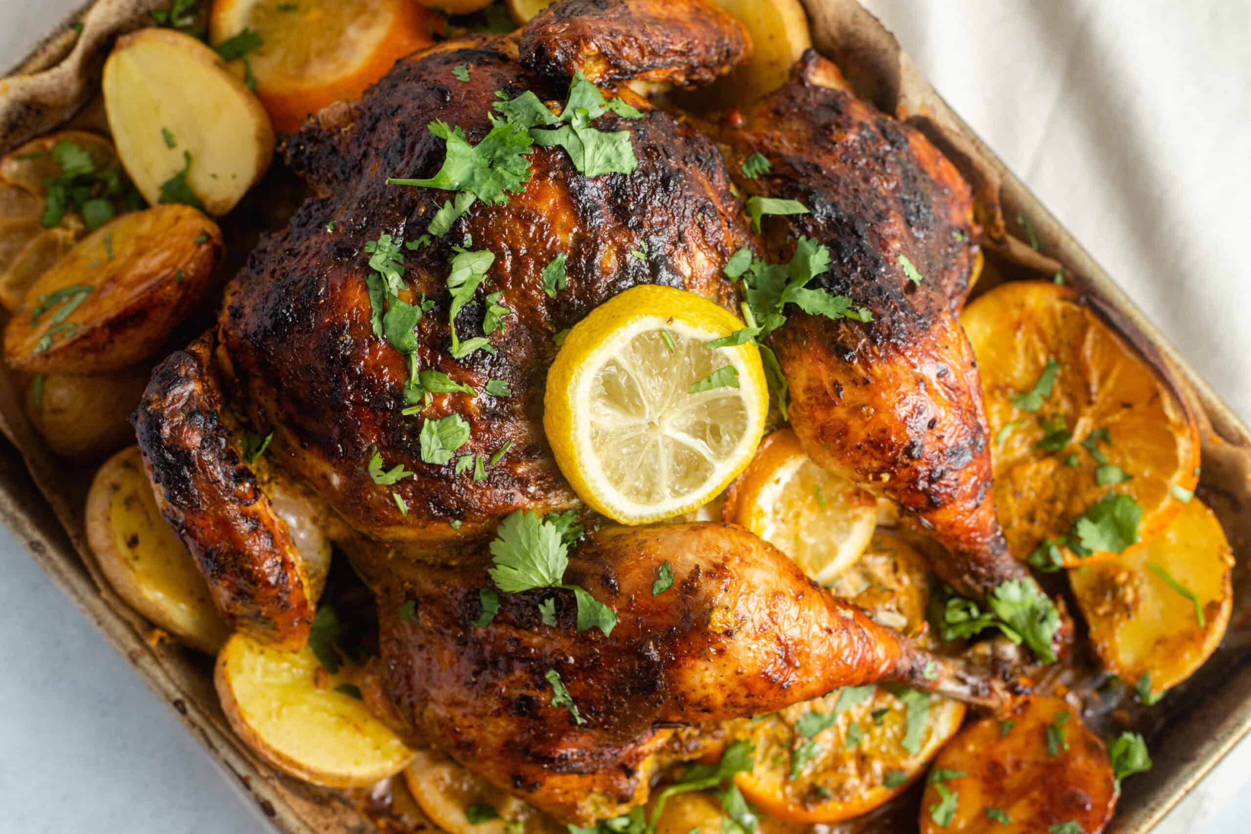 A baking sheet with a roast chicken topped with lemons sitting over a bed of potatoes.